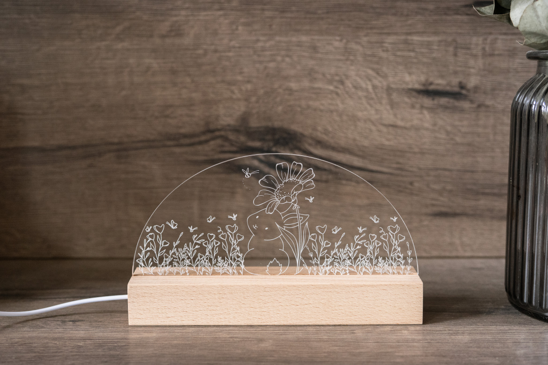 LED Osterlampe aus Acrylglas mit kleinem Hasen unter großer Blume in Wiese auf Holzsockel als Osterdeko