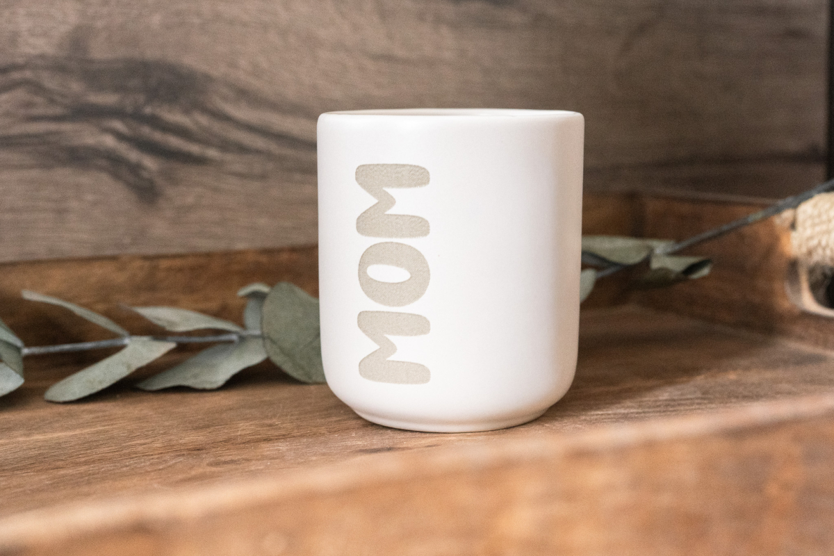 Matte weiße Keramiktasse mit vertikaler Gravur „MOM“ in moderner Schrift auf Holzuntergrund, Eukalyptus im Hintergrund.